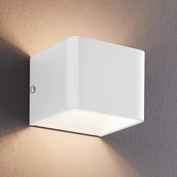 Biela nástenná lampa svieti na stenu. Lampa je štvorcového tvaru a pripevnená na stenu. Svetlo vychádza z jej spodnej časti.